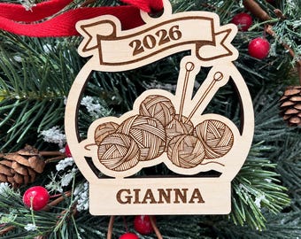 May include: A light-colored wooden ornament featuring a laser-engraved design of yarn balls and knitting needles. A banner at the top displays the year 2026, and a rectangular base at the bottom is engraved with the name GIANNA. A red ribbon is attached.