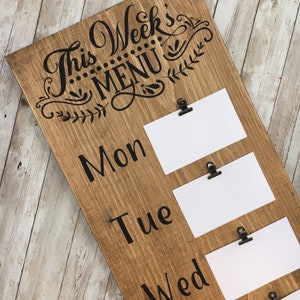 May include: A wooden menu board with the text "This Week's Menu" in black script lettering. The board has five spaces for cards with clips to hold them in place. The days of the week are labeled "Mon", "Tue", "Wed", "Thu", and "Fri" in black lettering.