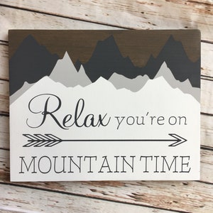 May include: Wooden sign with a mountain range design in shades of gray and white, topped with a brown border. The sign reads "Relax you're on MOUNTAIN TIME" with an arrow graphic.