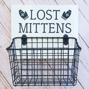 May include: A white sign with black text that reads "LOST MITTENS" with a black mitten graphic on each side. A black wire basket is attached to the sign.