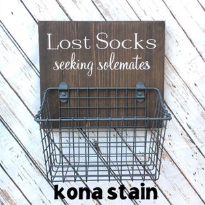May include: A dark brown wooden sign with white text that reads "Lost Socks seeking solemates". A gray wire basket is attached to the sign.