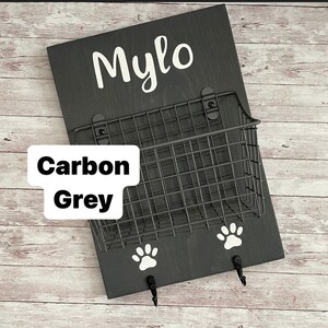 May include: A black wooden dog leash holder with the name "Mylo" written in white lettering. The holder has a wire basket and two hooks with paw print designs. The holder is labeled "Carbon Grey".