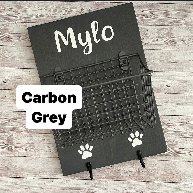May include: A black wooden dog leash holder with the name "Mylo" written in white lettering. The holder has a wire basket and two hooks with paw print designs. The holder is labeled "Carbon Grey".