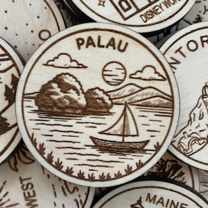 May include: A light brown wooden coaster with a laser-etched design. The coaster features the word "PALAU" at the top, a sailboat, mountains, clouds, and a sun. The design is etched into the wood.