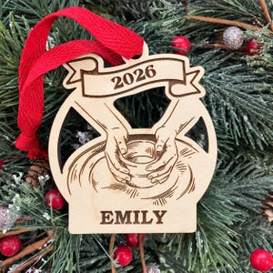May include: A light-colored wooden ornament featuring a laser-cut design of two hands shaping a clay pot on a wheel. A banner at the top displays the year 2026, and the name EMILY is engraved at the bottom. A bright red ribbon is attached for hanging.