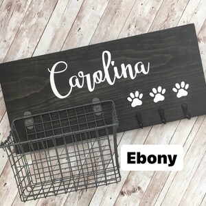 May include: Black wooden dog leash holder with the name "Carolina" in white lettering. The holder has three hooks and a wire basket. The wood is stained ebony.
