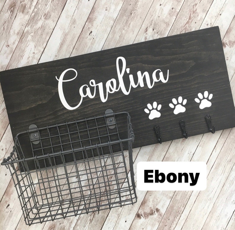 May include: Black wooden dog leash holder with the name "Carolina" in white lettering. The holder has three hooks and a wire basket. The wood is stained ebony.