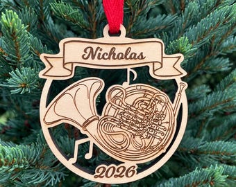 May include: A circular wooden ornament featuring a laser-cut French horn design. A banner at the top displays the name Nicholas, and the year 2026 is engraved at the bottom. The ornament is suspended by a bright red ribbon.