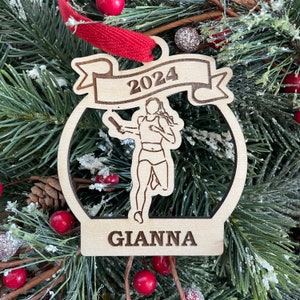 May include: A wooden Christmas ornament featuring a silhouette of a runner holding a baton. The ornament is decorated with the year "2024" and the name "Gianna".
