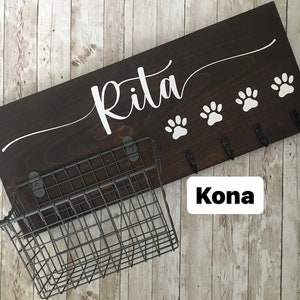 May include: Dark brown wooden dog leash rack with the name "Rita" written in white cursive lettering. The rack has four hooks and a wire basket underneath. The text "Kona" is written in white on a white rectangle.