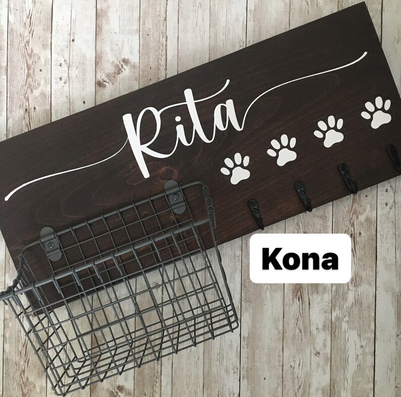 May include: Dark brown wooden dog leash rack with the name "Rita" written in white cursive lettering. The rack has four hooks and a wire basket underneath. The text "Kona" is written in white on a white rectangle.