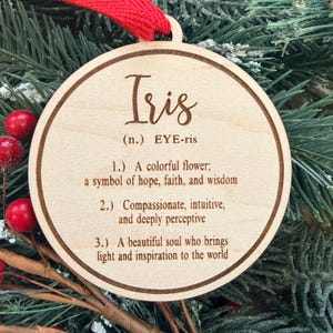 May include: A round, light-colored wooden ornament featuring a red ribbon loop at the top. The surface is laser-engraved with the name Iris, its phonetic pronunciation, and three definitions describing the name as a flower and a beautiful soul.