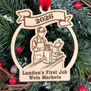 May include: A light-colored wooden ornament featuring an engraved illustration of a person in an apron packing a bag at a checkout counter. The banner at the top displays the year 2026, and the bottom text reads Landon's First Job Weis Markets.