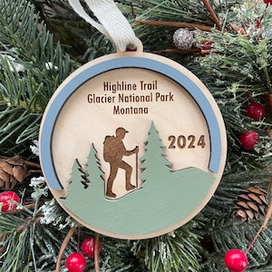 May include: A wooden Christmas ornament with a blue and white border. The ornament features a silhouette of a hiker on a trail with trees and the text "Highline Trail Glacier National Park Montana 2024".