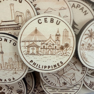 May include: A circular light-colored wooden disc featuring a laser-engraved illustration of a bridge, a church, and palm trees with the text CEBU PHILIPPINES centered on the surface. The design is part of a collection of similar wooden discs.