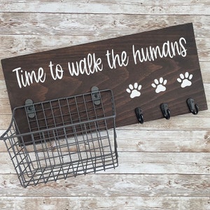 May include: A dark brown wooden sign with the text "Time to walk the humans" in white lettering. The sign has three black hooks and a white paw print design. A black metal wire basket is attached to the sign.