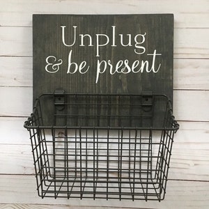 May include: A dark gray wooden sign with white text that reads "Unplug & be present". A black wire basket is attached to the bottom of the sign.