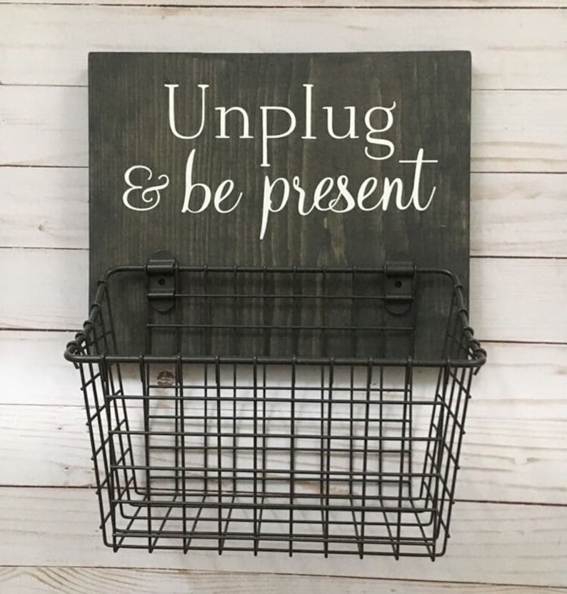May include: A dark gray wooden sign with white text that reads "Unplug & be present". A black wire basket is attached to the bottom of the sign.