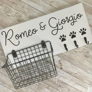May include: White wooden sign with black lettering that reads "Romeo & Giorgio" with three black paw prints and three black hooks. A gray wire basket is attached to the sign.
