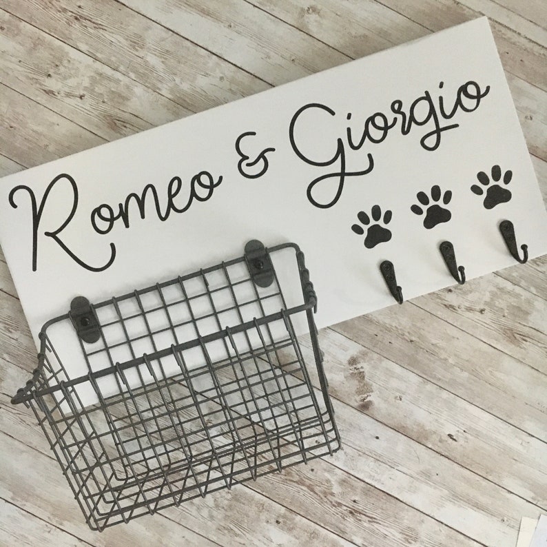 May include: White wooden sign with black lettering that reads "Romeo & Giorgio" with three black paw prints and three black hooks. A gray wire basket is attached to the sign.