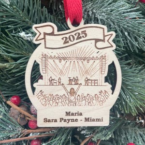 May include: Wooden Christmas ornament with a concert scene, featuring a stage, band, and crowd. The ornament has the year "2025" and the names "Maria Sara Payne - Miami" engraved on it. A red ribbon is attached for hanging.