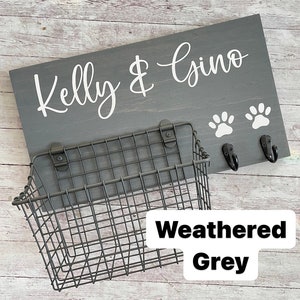 May include: A weathered grey wooden sign with the text "Kelly & Gino" in white lettering. Two white paw prints are below the text. A black metal wire basket is attached to the sign with two hooks.
