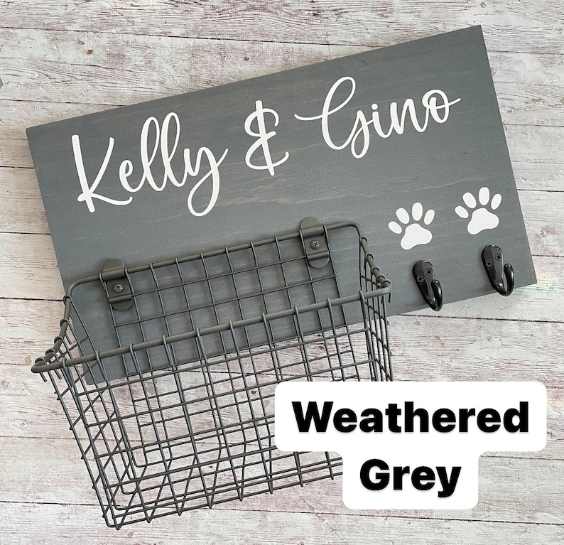 May include: A weathered grey wooden sign with the text "Kelly & Gino" in white lettering. Two white paw prints are below the text. A black metal wire basket is attached to the sign with two hooks.