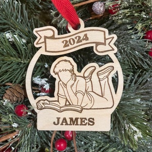 May include: A wooden Christmas ornament featuring a person reading a book, with the year 2024 and the name "JAMES" engraved on it.