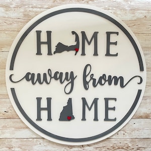 May include: A round wooden sign with a gray border and white background. The sign says "Home away from Home" with the states of Massachusetts and New Hampshire in the middle of the sign.