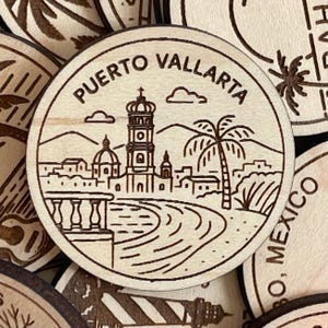 Puede incluir: Un recuerdo redondo de madera con un grabado de Puerto Vallarta, México. El diseño incluye una iglesia, una palmera y el texto "PUERTO VALLARTA". La imagen está rodeada de otros recuerdos de madera similares.