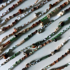 Puede incluir: Una colección de cordones trenzados coloridos con cuentas y dijes. Los cordones están hechos de diferentes materiales, incluyendo algodón, cáñamo y seda. Las cuentas están hechas de madera, vidrio y metal. Los dijes están hechos de metal y madera. Los cordones están dispuestos en un patrón aleatorio sobre un fondo blanco.