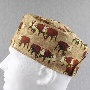 May include: A brown and white patterned cap featuring a repeating design of brown and white cows. The cap is worn on a white mannequin head. The cap is made of fabric and has a square shape.