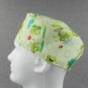 Pode incluir: Uma touca cirúrgica verde clara com um padrão divertido de sapos. A touca tem uma faixa branca e é decorada com sapos de desenho animado, corações e flores. Os sapos são retratados em várias poses, adicionando um toque lúdico ao design.