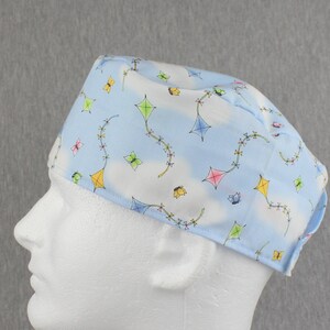 Puede incluir: Un gorro de quirófano azul claro con fondo blanco y un patrón de cometas y mariposas de colores.