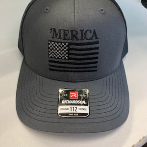 Könnte beinhalten: Graue Trucker-Kappe mit schwarzem Netzrücken. Die Vorderseite der Kappe zeigt eine schwarze gestickte amerikanische Flagge mit dem Wort "MERICA" darüber. Die Kappe hat auch ein schwarz-weißes Etikett mit der Aufschrift "Richardson" und "Original 112 Trucker One Size".