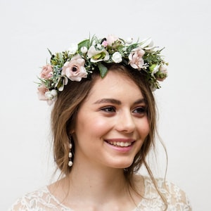 May include: A woman wearing a white lace dress with a floral crown on her head. The crown is made of pink and white flowers and greenery.