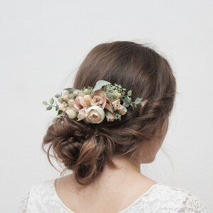 May include: A floral hair comb with blush pink and white flowers and greenery. The comb is worn in a woman's hair.