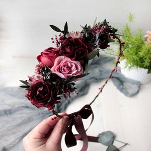 May include: A burgundy floral crown featuring pink roses and burgundy berries. The crown is crafted with a brown wire base and has a burgundy ribbon tied at the bottom.