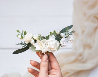 May include: A white floral hair comb with small white flowers and green leaves. The comb is held in a hand.