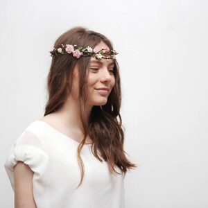 May include: A young woman with long brown hair wearing a white shirt and a floral crown with pink flowers.