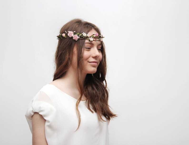 May include: A young woman with long brown hair wearing a white shirt and a floral crown with pink flowers.