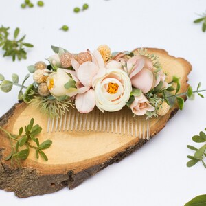 May include: A floral hair comb with blush pink and white flowers, greenery, and gold accents. The comb is displayed on a wooden slice.
