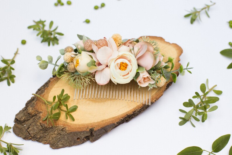 May include: A floral hair comb with blush pink and white flowers, greenery, and gold accents. The comb is displayed on a wooden slice.