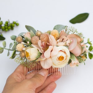 May include: A blush pink and cream floral hair comb with greenery and gold accents. The comb is held in a hand.