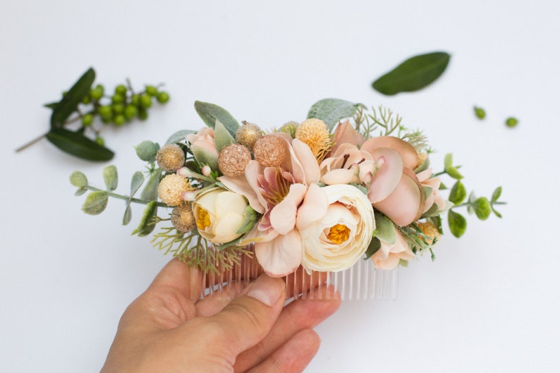 May include: A blush pink and cream floral hair comb with greenery and gold accents. The comb is held in a hand.