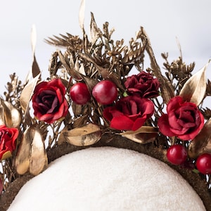 May include: A gold-toned floral crown with red roses and berries. The crown features gold leaves and intricate detailing, creating a regal and elegant aesthetic. The base is a neutral color, providing a contrast to the vibrant flowers.