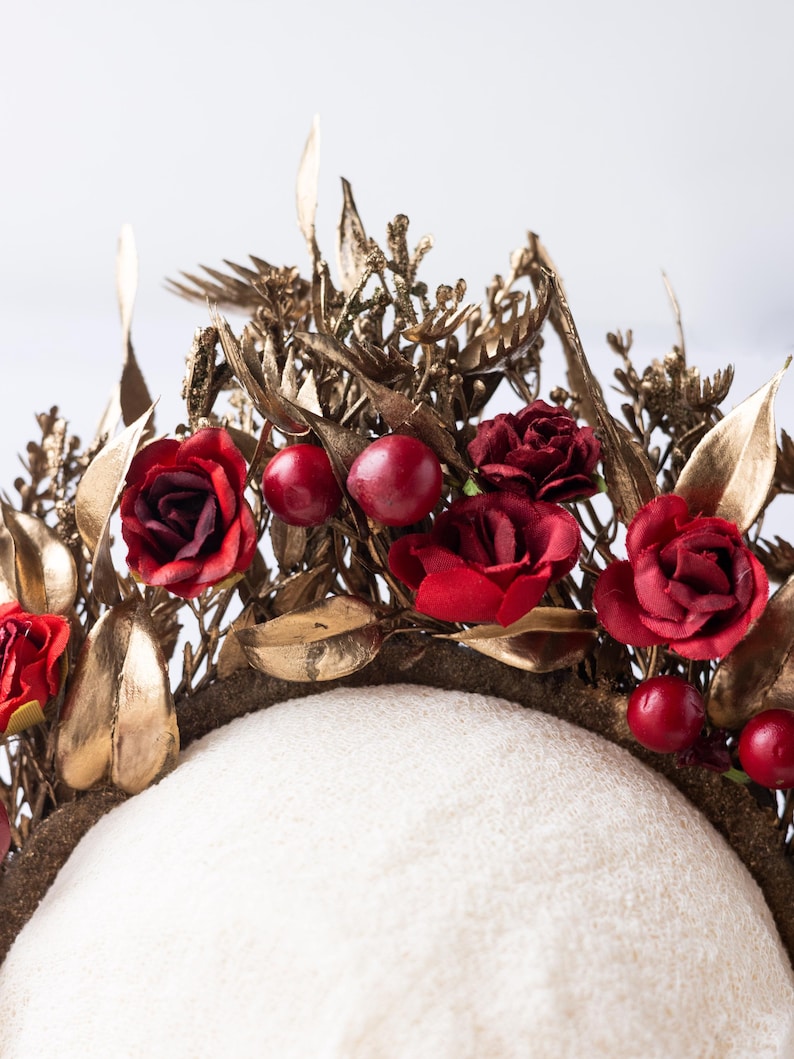 May include: A gold-toned floral crown with red roses and berries. The crown features gold leaves and intricate detailing, creating a regal and elegant aesthetic. The base is a neutral color, providing a contrast to the vibrant flowers.