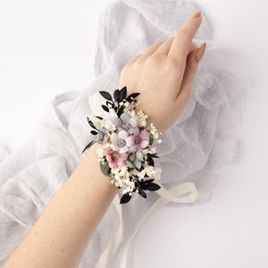 May include: A wrist corsage with white, pink, and blue flowers, black leaves, and white baby's breath. The corsage is tied with a white satin ribbon.