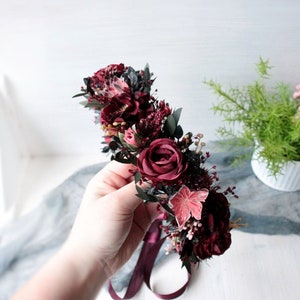 May include: A burgundy floral crown with a burgundy ribbon. The crown is made of silk flowers and greenery.