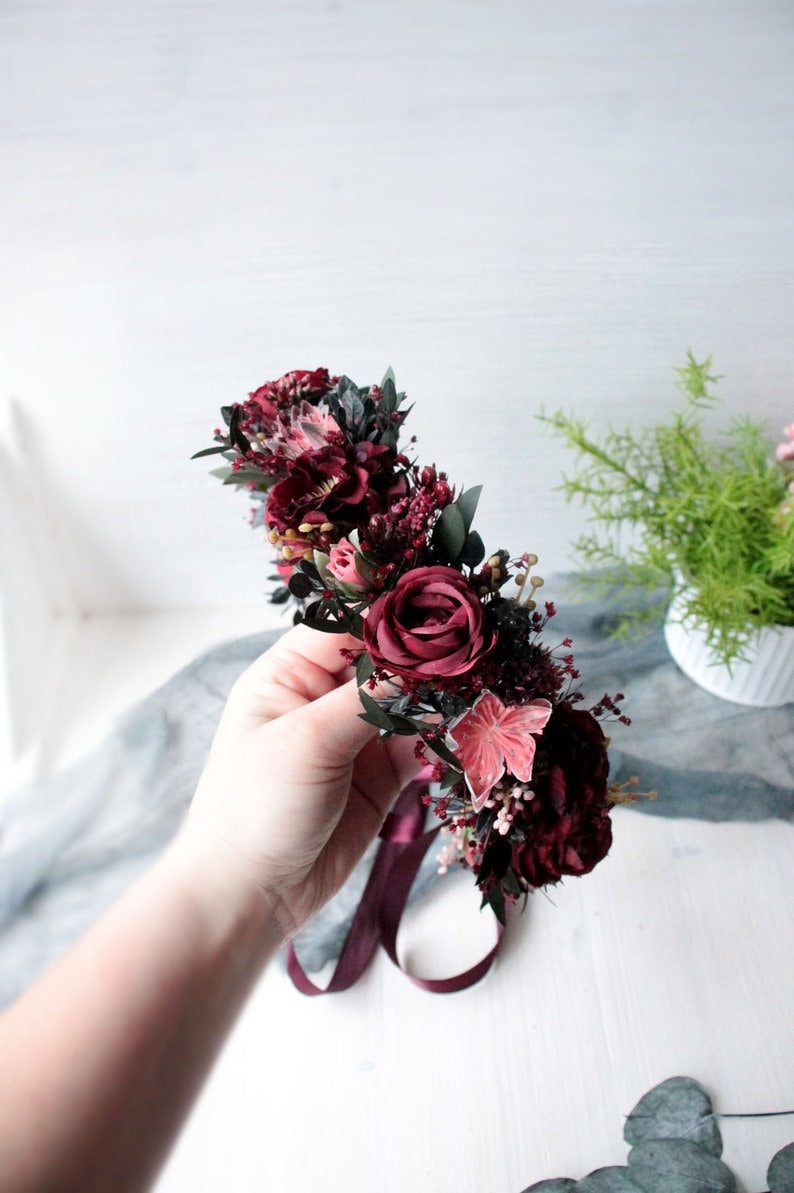 May include: A burgundy floral crown with a burgundy ribbon. The crown is made of silk flowers and greenery.